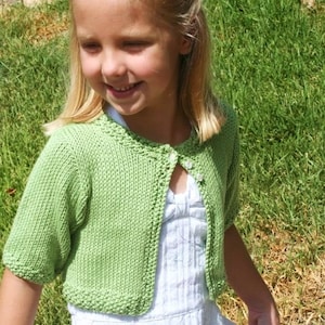 May include: A young girl wearing a green knitted cardigan with a white button closure over a white dress. She is standing in a grassy area.