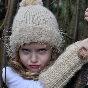 May include: A young girl wearing a white knitted hat with a fluffy pom pom and matching knitted arm warmers. The hat has a fluffy trim around the brim. The girl is looking at the camera with a serious expression.