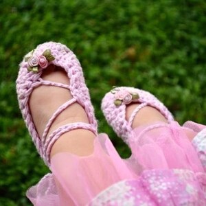 Könnte beinhalten: Rosa gehäkelte Babyschuhe mit einer Blumenverzierung. Die Schuhe haben ein Kreuzriemen-Design und sind mit einem Band gebunden.