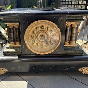 May include: A black mantel clock with a gold-toned circular face featuring black Arabic numerals. The clock case includes decorative gold-colored columns, lion head accents with rings on the sides, and ornate gold feet with intricate scrollwork.