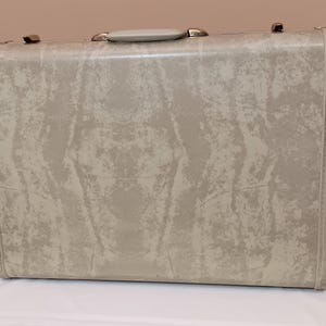May include: A vintage suitcase in a light gray color with a textured, mottled pattern. The suitcase has a handle and metal clasps. The overall design is rectangular, with rounded corners and a stitched edge.