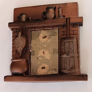 May include: A bronze-colored decorative fireplace with a rustic design. It features a brick facade, a wooden mantel with miniature jugs and a candle, and a central panel with sunflower artwork. A small pot and bellows are also included.