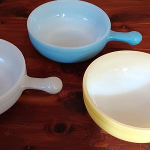 May include: Three colorful, round bowls with handles. One is white, one is light blue, and the other is yellow. The bowls are made of a glossy material and are set on a wooden surface. They are likely for serving soup or other dishes.