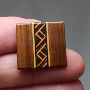 May include: A small, square wooden object with a decorative inlaid design. The object is primarily brown with a central band of red, gold, and brown geometric patterns. The item is held in a person's hand.