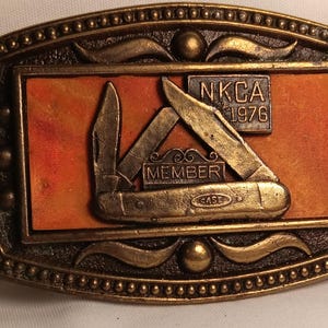 May include: A rectangular, bronze-colored belt buckle with a knife design. The buckle features the text "NKCA 1976" and "MEMBER" with a "CASE" knife. The background is orange.