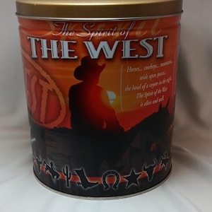 May include: A cylindrical tin with a gold lid and a design featuring a cowboy on horseback silhouetted against a sunset. The text "The Spirit of the West" is prominently displayed, with additional text about cowboys and open spaces.