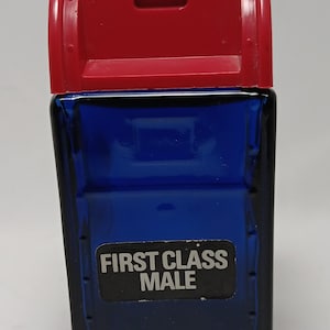 May include: A rectangular blue glass bottle shaped like a mailbox with a red plastic lid. The front of the bottle features a black rectangular label with the white text FIRST CLASS MALE printed in capital letters. The design includes a slot.
