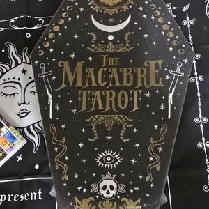 May include: A black coffin-shaped tarot card box with gold lettering and illustrations. The box reads "The Macabre Tarot" and features images of a bat, moon, snakes, and a skull. The artist's name, Sam Rook, is printed at the bottom.