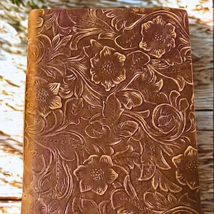 May include: A brown leather journal with a floral embossed design. The cover features a detailed pattern of flowers and leaves, creating a textured appearance. The journal is rectangular and appears to be closed.
