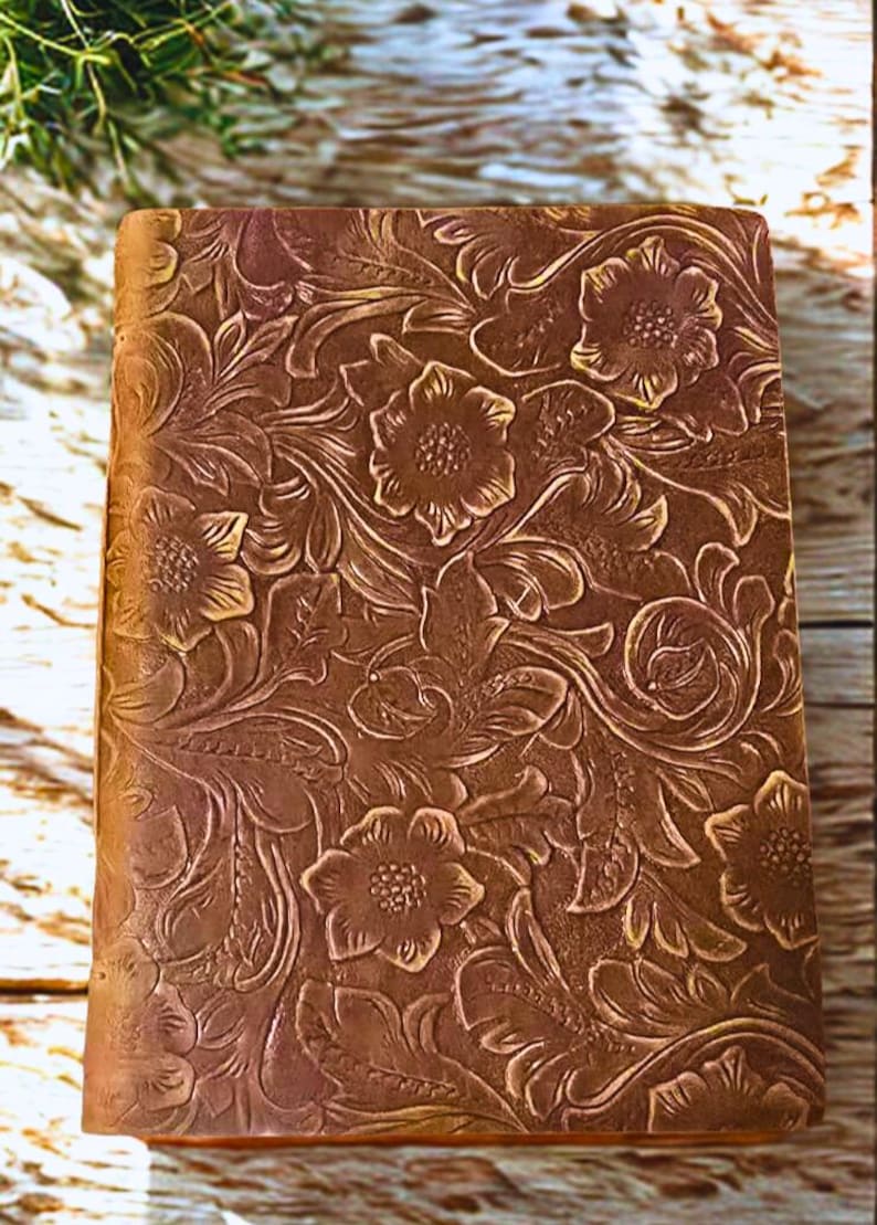 May include: A brown leather journal with a floral embossed design. The cover features a detailed pattern of flowers and leaves, creating a textured appearance. The journal is rectangular and appears to be closed.