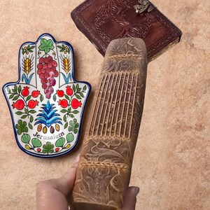 May include: A hand-shaped ceramic dish with a Jerusalem design, featuring colorful floral and fruit motifs. A carved wooden book and a leather-bound journal with a decorative clasp are also visible.