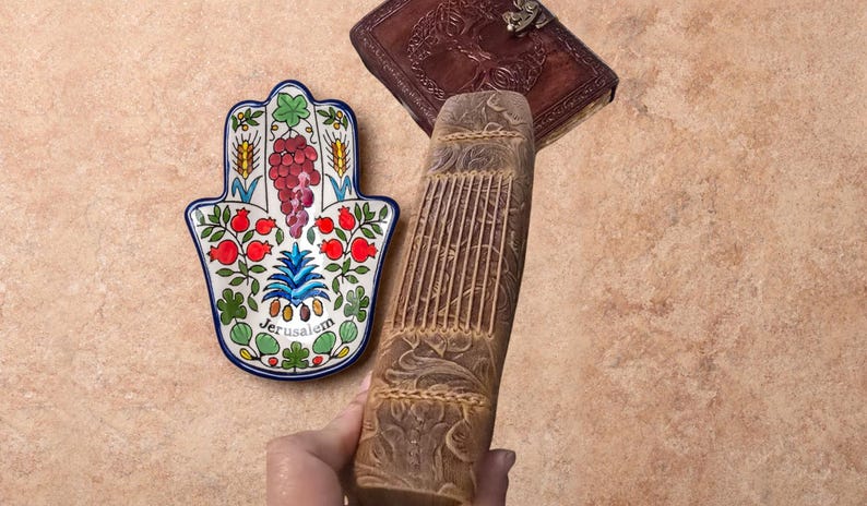 May include: A hand-shaped ceramic dish with a Jerusalem design, featuring colorful floral and fruit motifs. A carved wooden book and a leather-bound journal with a decorative clasp are also visible.