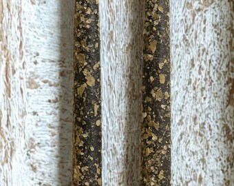 May include: Two black taper candles with a textured surface, speckled with gold-colored flakes. The candles have a rustic, handmade appearance, perfect for home decor or special occasions.