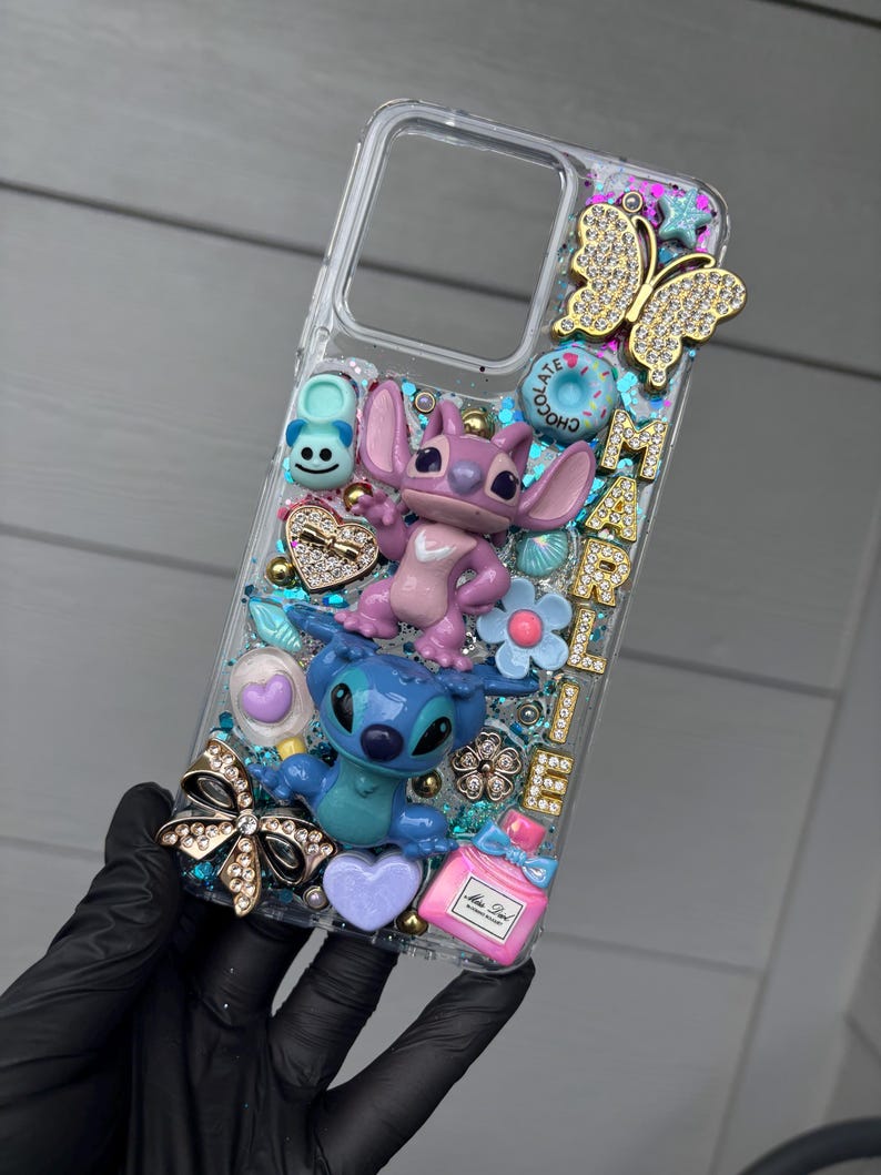 May include: Clear phone case decorated with Stitch and Angel figurines, a gold butterfly, and various embellishments. The case features the name "MARIEL" in gold letters, along with hearts, flowers, and a perfume bottle.