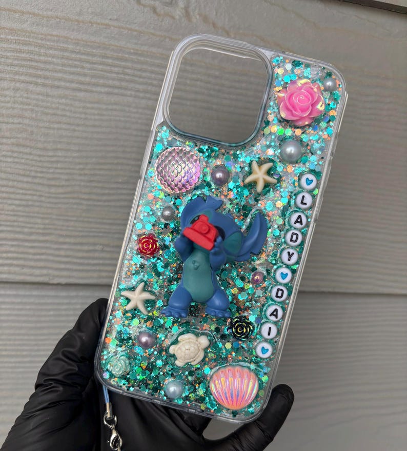 May include: Clear phone case with a glittery teal background, featuring a blue cartoon character, pink and red roses, pearls, and starfish. The name "LADY DAI" is spelled out in white letters.
