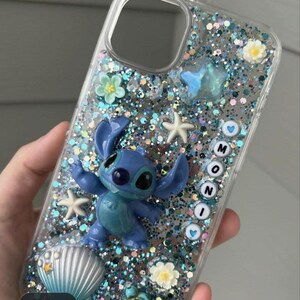 May include: Clear phone case with a blue Stitch figure, glitter, and decorative elements. The case features a blue Stitch figurine, starfish, flowers, and the word "MONI" spelled out with letter beads. The case is filled with blue and silver glitter.