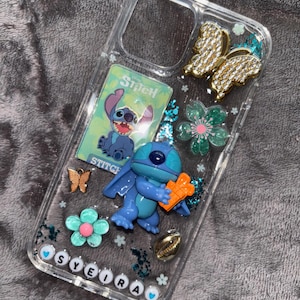 May include: Clear phone case decorated with a blue Stitch figure, a Stitch card, gold and bronze butterflies, and flower accents. The name "SYEIRA" is spelled out in white letter beads at the bottom of the case.