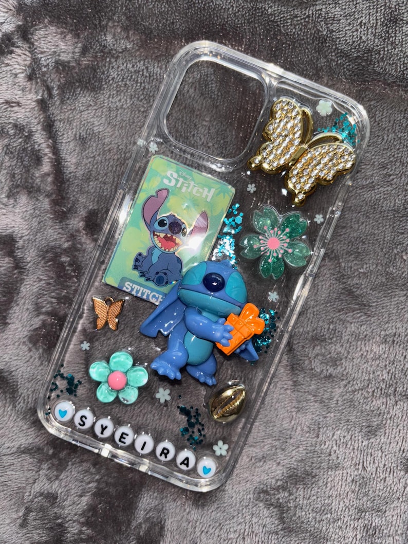May include: Clear phone case decorated with a blue Stitch figure, a Stitch card, gold and bronze butterflies, and flower accents. The name "SYEIRA" is spelled out in white letter beads at the bottom of the case.