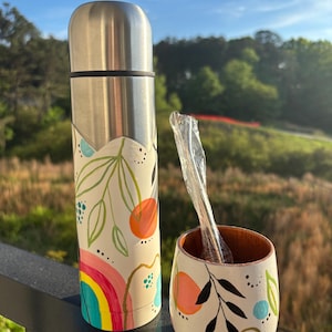 May include: A stainless steel thermos and matching cup set. The thermos and cup are painted with a cream-colored base and feature a colorful design of a rainbow, orange fruit, and green leaves. A clear straw is in the cup.