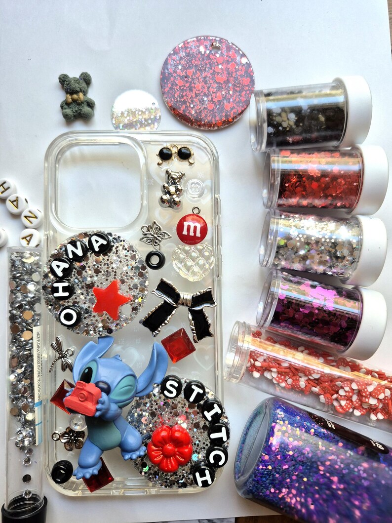 May include: Clear phone case decorated with glitter, charms, and a Stitch figurine. The case features the word "OHANA" and "STITCH" in black letters, along with a red star, bow, and flower. Several containers of glitter and beads are also visible.