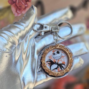 May include: A circular keychain featuring a central illustration of a skeletal character with a wide grin and a black pinstriped suit. The image is set within a decorative gold-toned scalloped frame, accented by orange paint splatters on a white background.