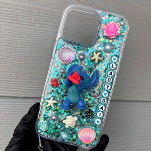 May include: A clear phone case filled with teal and blue glitter, featuring a blue character figurine holding a red camera. The case is decorated with a pink rose, white starfish, a turtle, a seashell, pearls, and beads spelling out LADY with hearts.