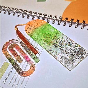 May include: A rectangular resin bookmark with an orange and green gradient filled with silver glitter, featuring a matching orange tassel and a separate oval-shaped resin paperclip with a similar glittery orange and green finish.