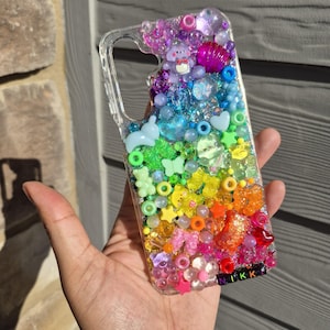 Puede incluir: Una funda de teléfono transparente decorada con una densa disposición de cuentas, dijes y lentejuelas de colores en un degradado de arcoíris. Los adornos incluyen pequeñas figuras de animales, estrellas, corazones y flores. El nombre NIKKI está escrito con cuentas de letras negras.
