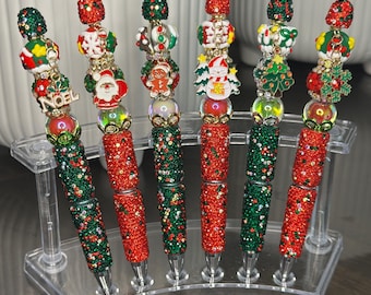 May include: A collection of Christmas-themed pens. Each pen features a red and green jeweled barrel, topped with decorative beads and charms. Charms include a Santa, snowman, Christmas tree, and snowflake. The pens are displayed in a clear acrylic holder.