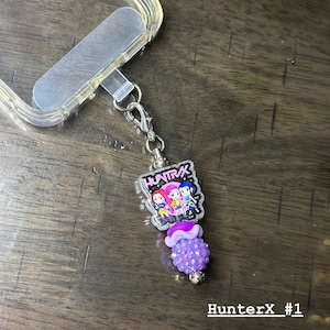 May include: A clear phone case with a silver-colored charm. The charm features a cartoon image with the text "HUNTRX" and a purple beaded accent. The charm is attached to the phone case with a silver-colored clasp. The text "HunterX_#1" is at the bottom.