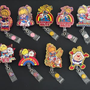 May include: A collection of Rainbow Brite character badge reels. Each reel features a colorful, glittery design with a Rainbow Brite character and the words "Rainbow Brite" or "Color Kids". The reels have a clear plastic strap and a metal clip.
