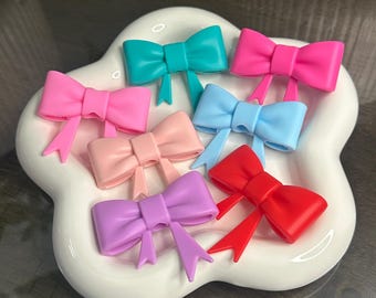 May include: A collection of colorful bow-shaped hair accessories in shades of pink, teal, blue, red, and purple. The bows are arranged on a white, cloud-shaped dish. The bows have a glossy finish and are likely made of plastic or resin.