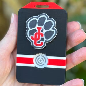 May include: A rectangular black plastic badge holder featuring a red top edge with a slot. The center displays a black paw print emblem with a red and white JC logo. Below, a horizontal red stripe crosses the body with a circular white icon.