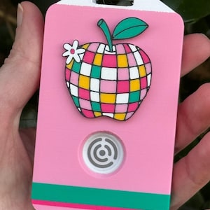 May include: A pink luggage tag with a colorful apple design. The apple is made up of squares of yellow, pink, green, and white. A white flower is next to the apple. The tag has a white top and a green stripe at the bottom.