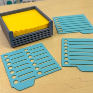 May include: A gray square holder containing a stack of bright yellow square sticky notes, accompanied by two light blue plastic stencils featuring horizontal lines and circular bullet points for organizing tasks on paper notes.