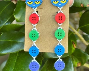 Könnte beinhalten: Hängeohrringe mit einem blauen Katzenkopf mit gelben Augen, verbunden mit einer Reihe roter, grüner, blauer und violetter Knöpfe. Die Ohrringe sind an einem braunen Etikett befestigt, vor einem grünen, blättrigen Hintergrund.