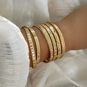 May include: A collection of gold bangles with various engraved patterns, including geometric shapes, stars, and floral designs. The bangles are stacked on a wrist, showcasing their different textures and styles. The jewelry is set against a white background.