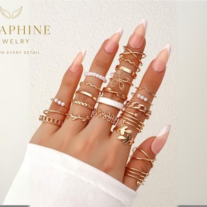 Könnte beinhalten: Eine Hand, die eine Sammlung goldfarbener Ringe zeigt. Die Ringe weisen verschiedene Designs auf, darunter Perlen, Kettenglieder und florale Muster. Das Bild enthält auch den Text "SERAPHINE JEWELRY" und "GRACE IN EVERY DETAIL".