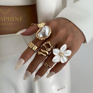 Op de afbeelding: Een hand toont een verzameling goudkleurige ringen, waaronder een grote parelring, een getextureerde bandring en een bloemvormige ring met een wit centrum. De ringen staan tegen een witte achtergrond.