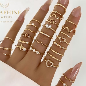 Può includere: Una collezione di anelli dorati in vari design, tra cui cuori, onde e fasce strutturate. Alcuni anelli presentano piccole gemme trasparenti. Gli anelli sono esposti su dita, mettendo in risalto i dettagli dei gioielli. Il marchio "SERAPHINE JEWELRY" è visibile.