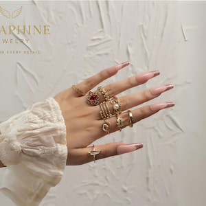 Puede incluir: Una mano que muestra varios anillos de oro, incluido un anillo con una piedra preciosa roja, un anillo de perlas y varios anillos de banda. Las uñas son largas y están pintadas con un diseño rosa y dorado. La muñeca está adornada con un puño de encaje blanco. La imagen incluye el texto "SERAPHINE JEWELRY".