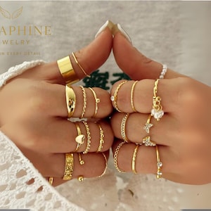 Può includere: Primo piano di mani adornate da anelli d'oro. I modelli includono fasce lisce, intrecciate e anelli con piccoli cuori e abbellimenti. Alcuni anelli presentano piccole gemme e perle. Il testo "SERAPHINE JEWELRY, GRACE IN EVERY DETAIL" è visibile.