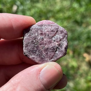 May include: A raw, hexagonal-shaped gemstone with a deep pinkish-purple hue. The surface of the stone displays a natural, crystalline texture with visible internal fractures, opaque sections, and small dark mineral inclusions throughout the structure.