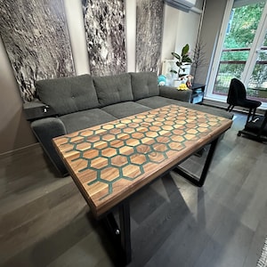Puede incluir: Una mesa de centro rectangular con una tapa de madera con un diseño de panal de abeja con incrustaciones de resina verde. La mesa tiene una estructura de metal negro y está situada en un salón.