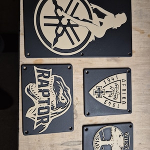 May include: A set of black rectangular plaques with beige designs. The plaques display a pin-up girl, a dinosaur with the word "RAPTOR", a military emblem with the text "AFRIKA 1941-1943", and a tree design with the word "WIESA".