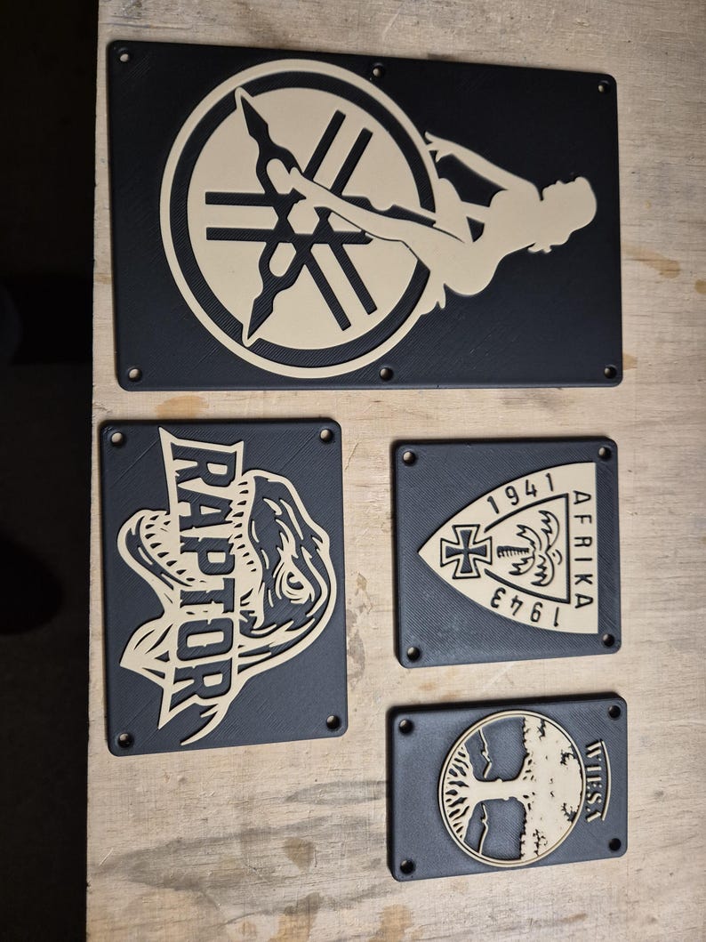 May include: A set of black rectangular plaques with beige designs. The plaques display a pin-up girl, a dinosaur with the word "RAPTOR", a military emblem with the text "AFRIKA 1941-1943", and a tree design with the word "WIESA".