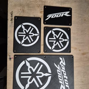 May include: A selection of black rectangular plates with white graphics. The plates display the Yamaha logo and the text "700R" and "Raptor 700R". These plates are likely for a motorcycle or ATV.