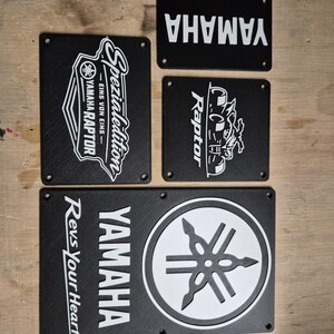 May include: A collection of black rectangular Yamaha signs with white text and logos. One sign features "Yamaha Raptor" with a graphic. Another says "Yamaha Spezial Edition". The bottom sign says "Yamaha Revs Your Heart".