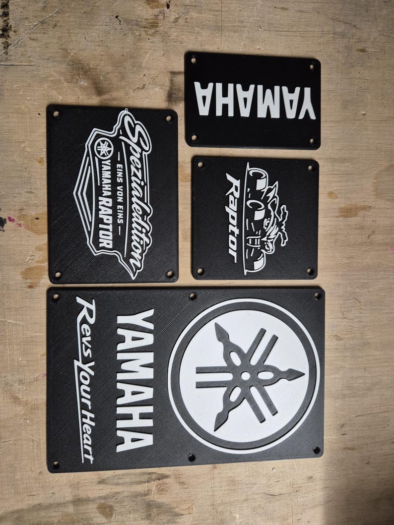 May include: A collection of black rectangular Yamaha signs with white text and logos. One sign features "Yamaha Raptor" with a graphic. Another says "Yamaha Spezial Edition". The bottom sign says "Yamaha Revs Your Heart".