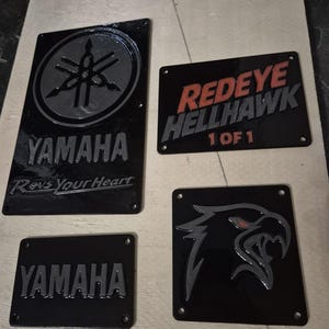 May include: Four black metal plaques with embossed designs. One plaque features the Yamaha logo and text, another reads "Redeye Hellhawk 1 of 1", and the others have stylized graphics. All plaques have mounting holes.